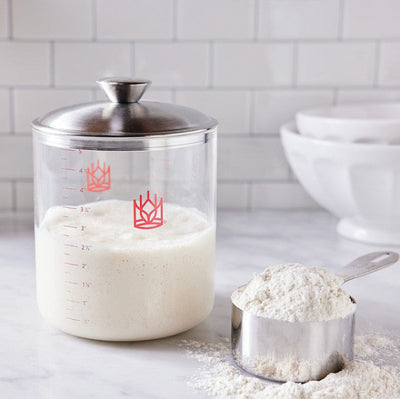 Glass sourdough container with sourdough starter and flour in measuring cup on a kitchen counter